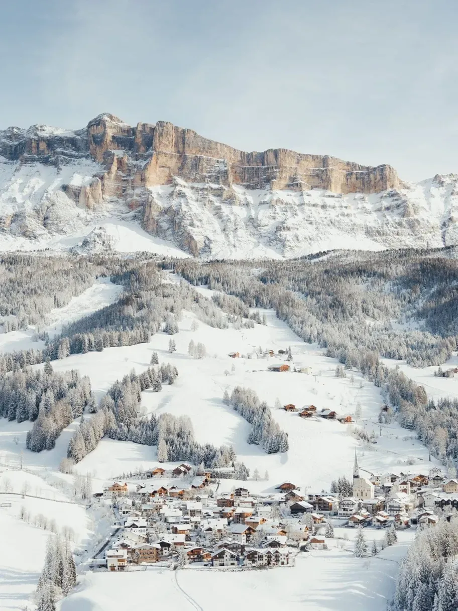 Paesaggio in Alta Badia, Dolomiti: villaggio innevato ai piedi di maestose cime rocciose, boschi innevati e chalet alpini sotto un cielo azzurro.