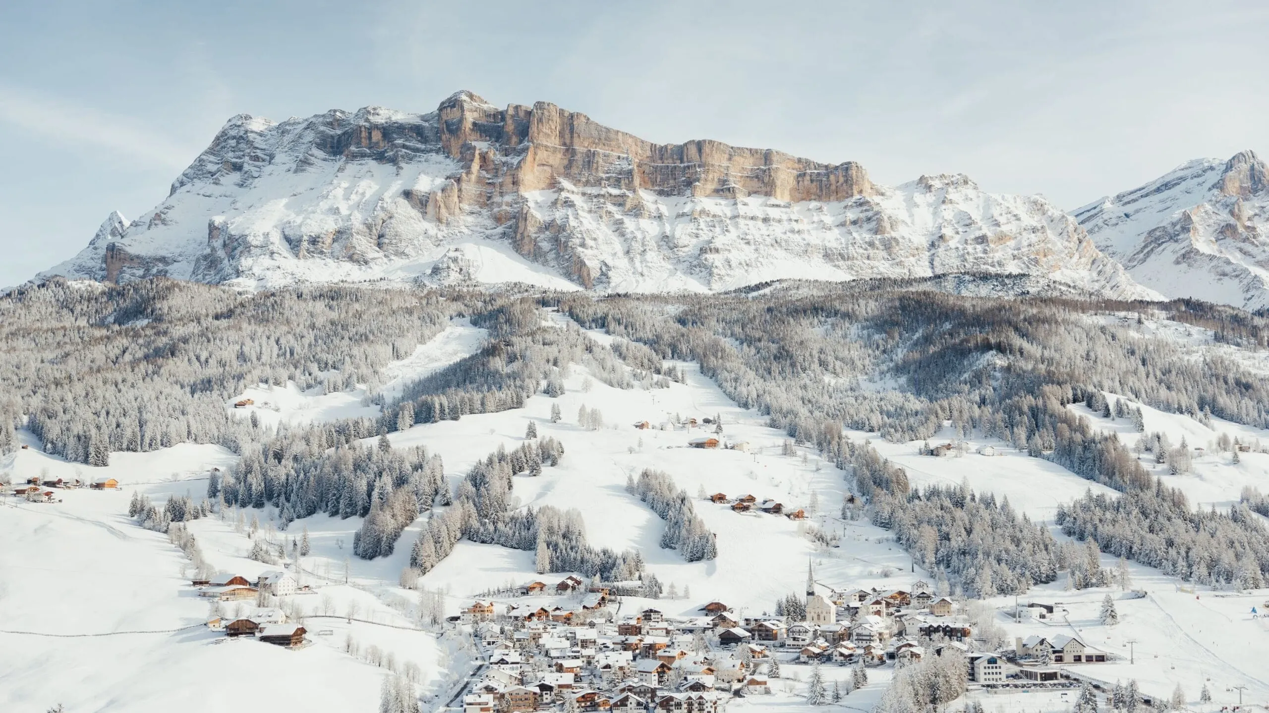 Paesaggio in Alta Badia, Dolomiti: villaggio innevato ai piedi di maestose cime rocciose, boschi innevati e chalet alpini sotto un cielo azzurro.