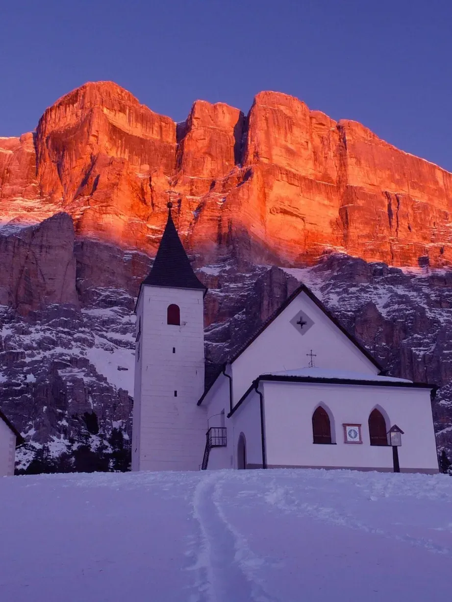 Chiesa bianca con campanile, edificio annesso a sinistra, terreno innevato; Dolomiti al tramonto con rocce dorate e cielo azzurro.
