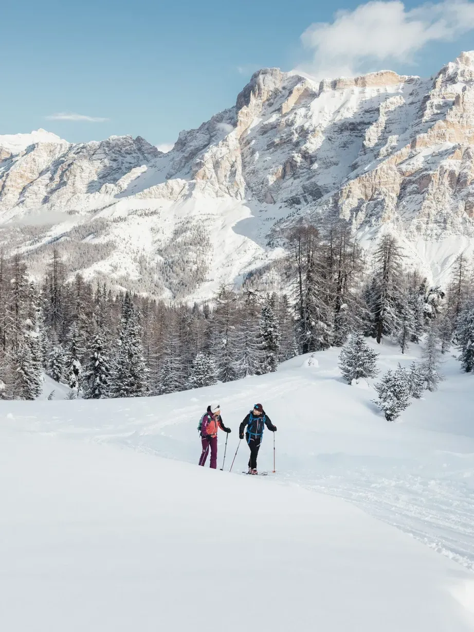 Due persone in sci di fondo lungo un sentiero innevato tra pini, con Dolomiti di Alta Badia innevate sullo sfondo e cielo azzurro.