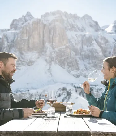 Due persone sedute a tavola di legno all'aperto tra le Dolomiti di Alta Badia, innevate; brindano con bicchieri di vino e gustano piatti, cielo sereno.
