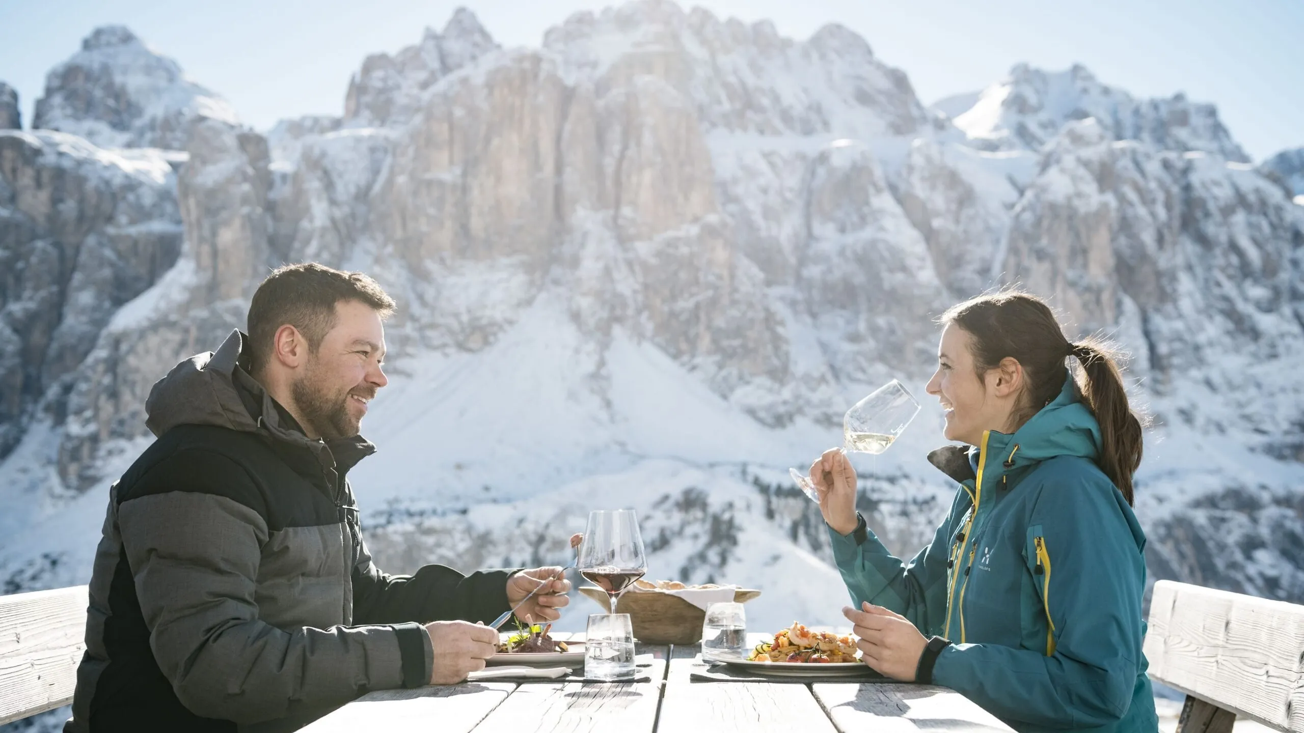 Due persone sedute a tavola di legno all'aperto tra le Dolomiti di Alta Badia, innevate; brindano con bicchieri di vino e gustano piatti, cielo sereno.