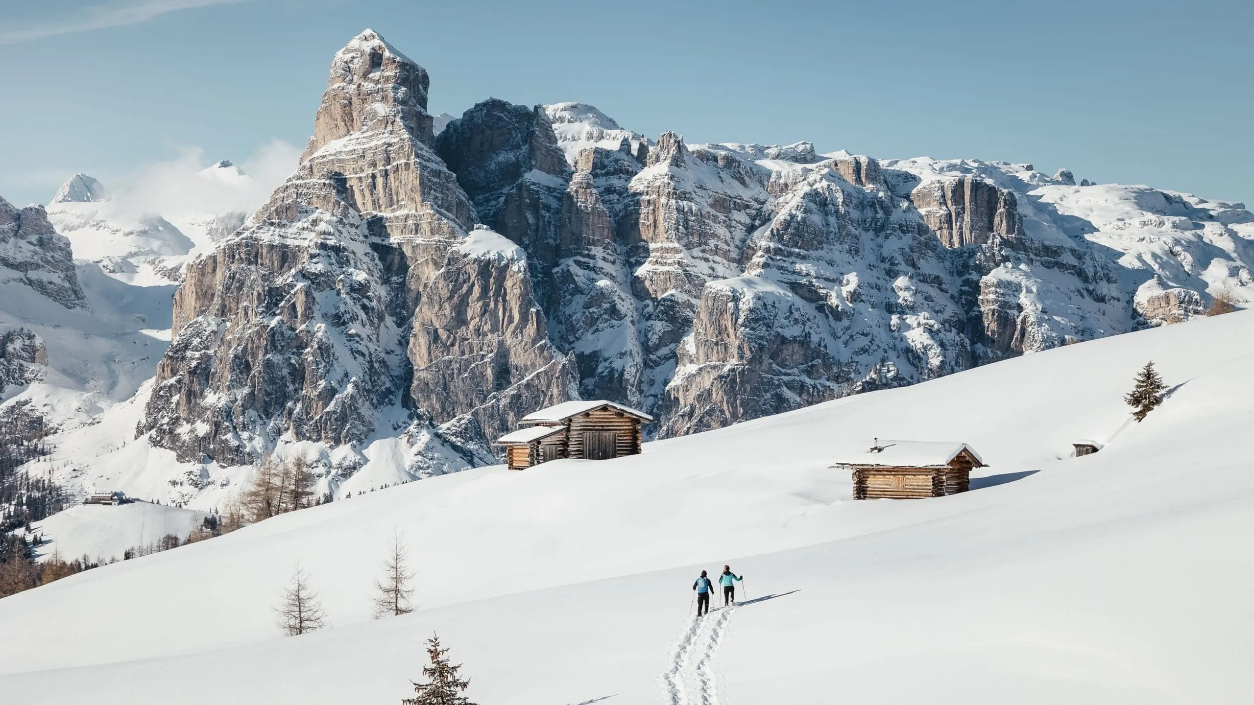 Due persone camminano sulla neve con bastoncini tra baite di legno nelle Dolomiti, Alta Badia, con picchi sullo sfondo e cielo azzurro.