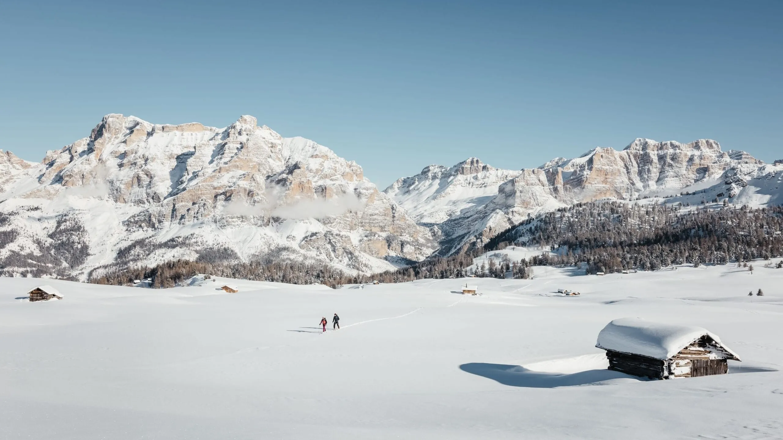 Paesaggio innevato nelle Dolomiti, Alta Badia: vaste vallate, alte montagne, due sciatori e piccole baite di legno, cielo sereno.