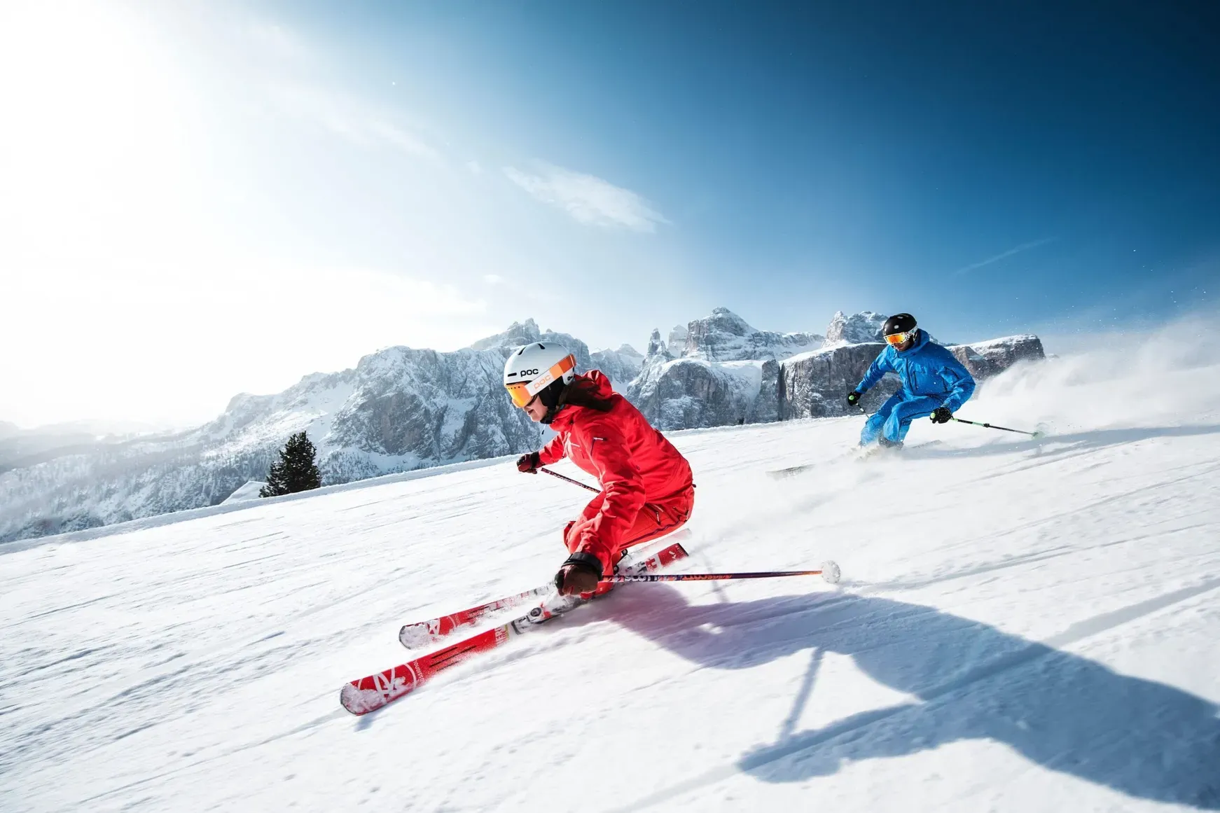 Two skiers, one in red and one in blue, carve a snowy slope with Dolomite peaks beneath a clear blue sky in Alta Badia, Dolomites.