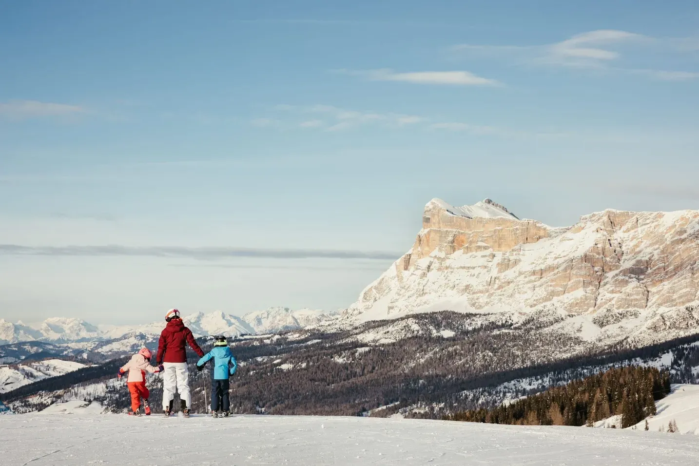 Three people, an adult and two children, in colorful winter gear on a snowy slope in Alta Badia Dolomites, with blue sky and rugged peaks behind.