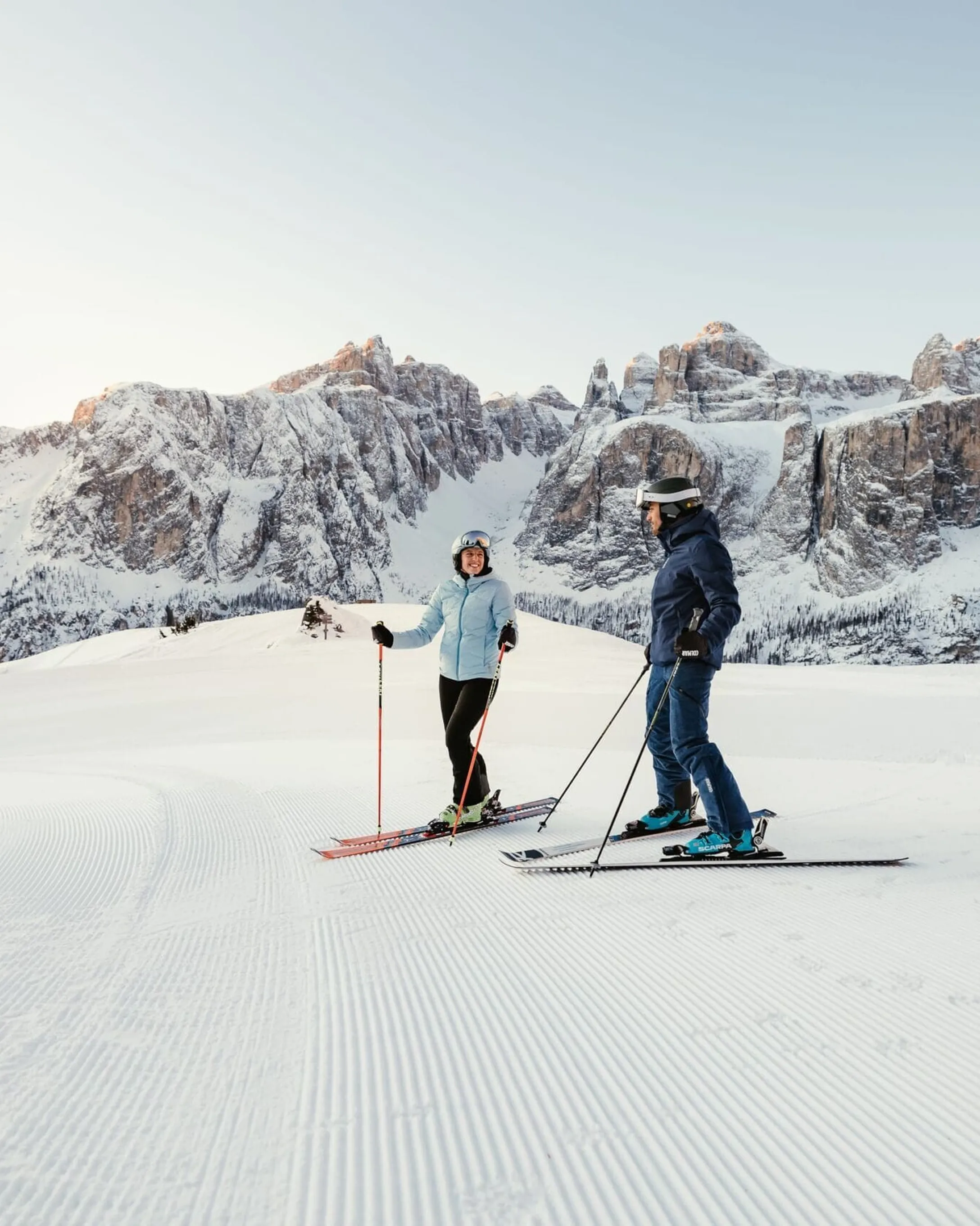 Two skiers on a groomed slope in Alta Badia, Dolomites, with snow-covered peaks rising in the background.
