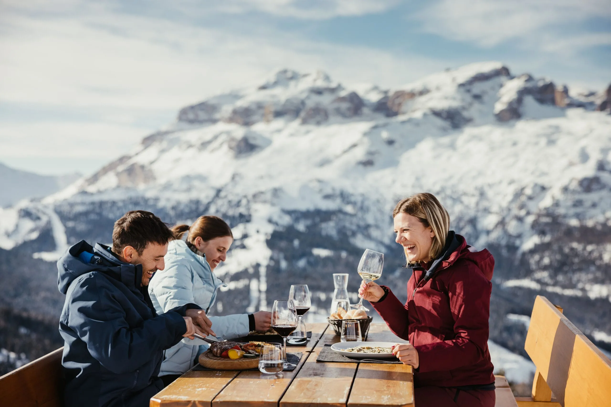 Three people in winter jackets dine outdoors at a wooden table in Alta Badia, Dolomites, with wine glasses, plates, and snow-covered peaks in the background.