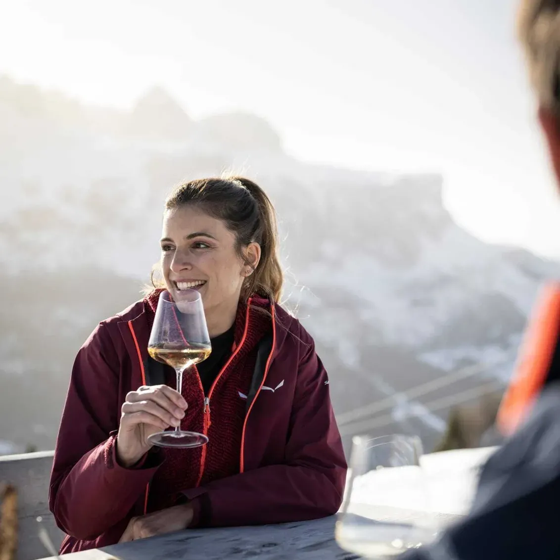 Persona seduta all'aperto ad Alta Badia, Dolomiti, sorride mentre brinda con un calice di vino; tavolo con cibo davanti e montagne innevate sullo sfondo.