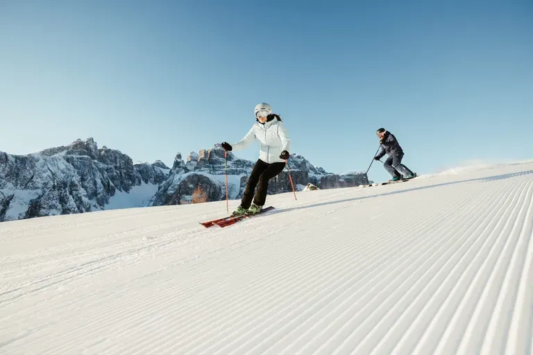 Two skiers descend a groomed slope in Alta Badia, Dolomites, with parallel grooves in the snow and rugged peaks under a clear blue sky.