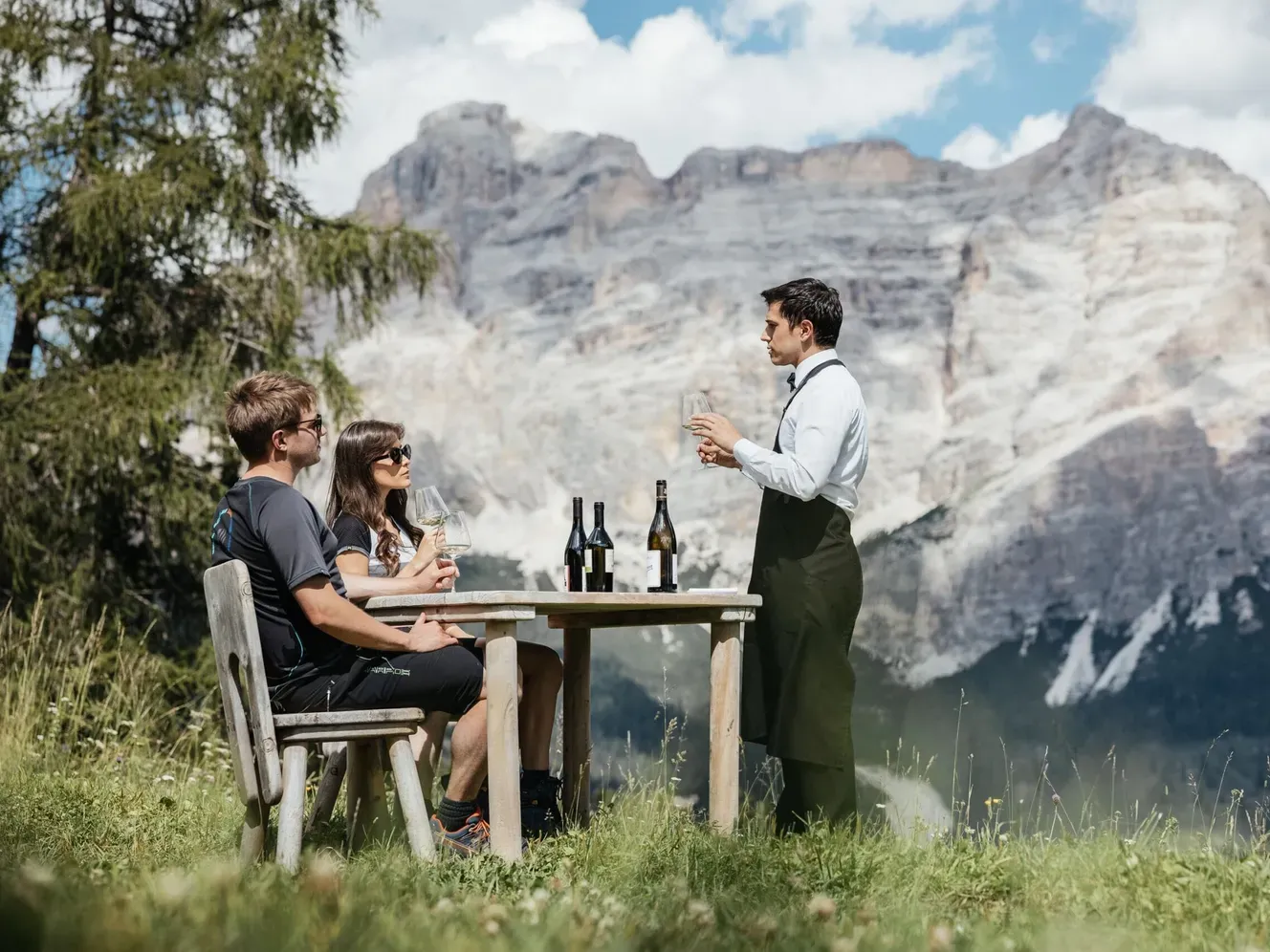 Tre persone all'aperto nelle Dolomiti dell'Alta Badia: due ospiti sorseggiano vino al tavolo di legno, mentre un addetto al servizio li serve.