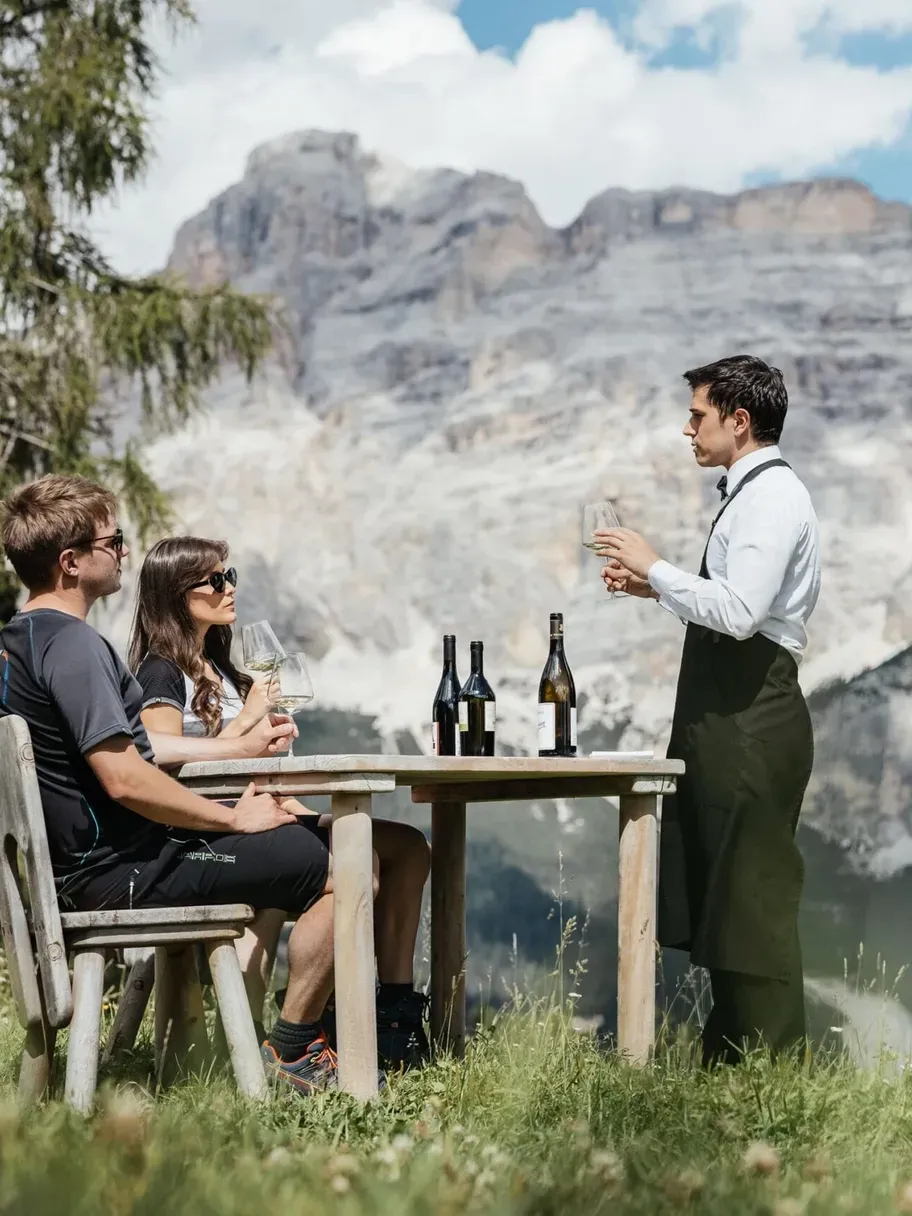 Three people sit at a wooden table on grassy slope in Alta Badia, Dolomites; a waiter pours wine, bottles on the table, with rugged mountain backdrop.