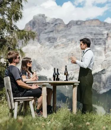 Three people sit at a wooden table on grassy slope in Alta Badia, Dolomites; a waiter pours wine, bottles on the table, with rugged mountain backdrop.