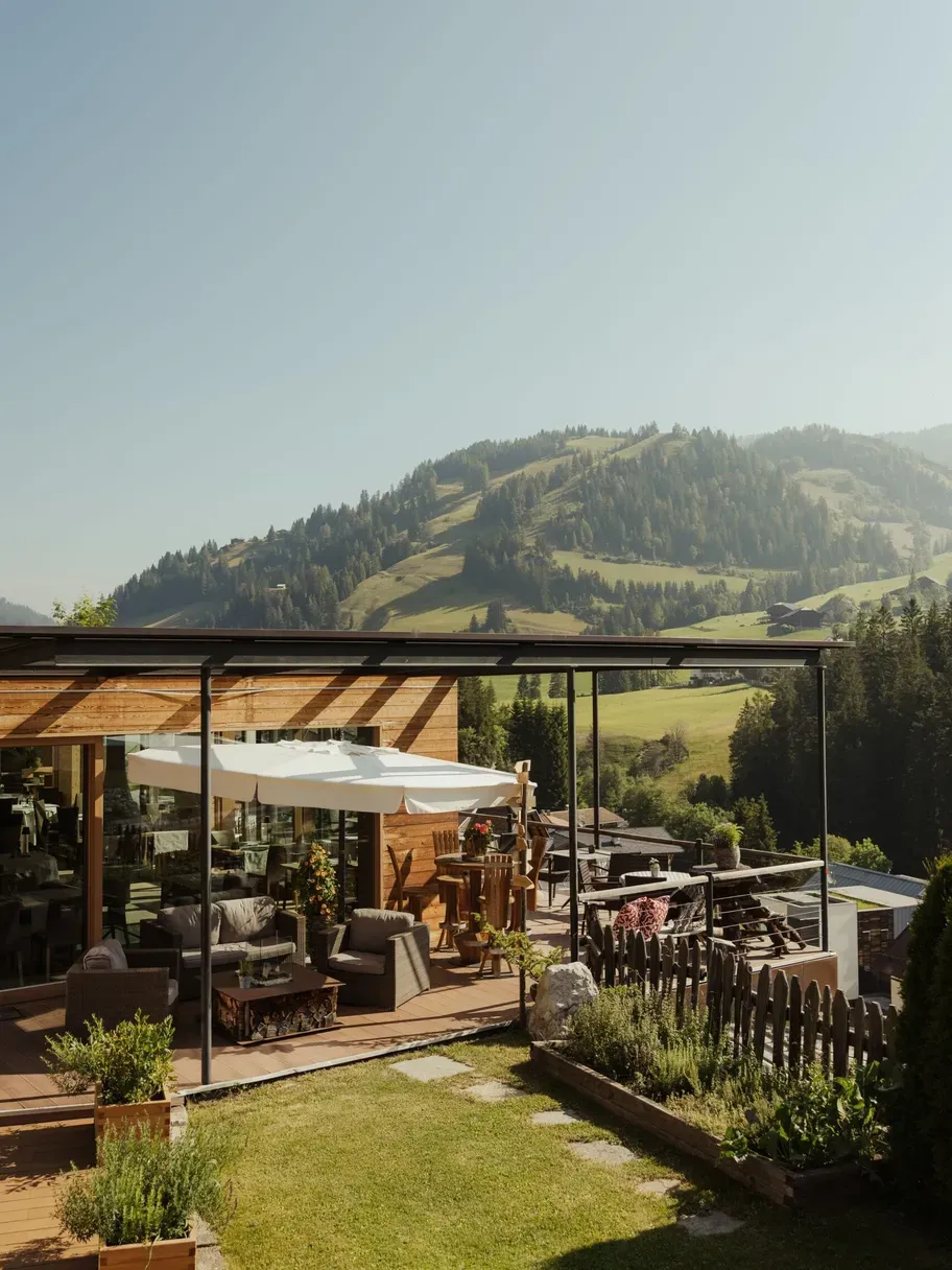 Hotel Gran Ander terrace in Alta Badia, Dolomites: glass-walled wooden deck with lounge seating, white umbrellas, and a mountain valley view.