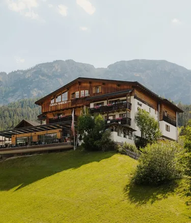 Chalet-style hotel on a grassy hillside in Alta Badia, Dolomites, with wooden balconies and a terrace, set against forested mountains.