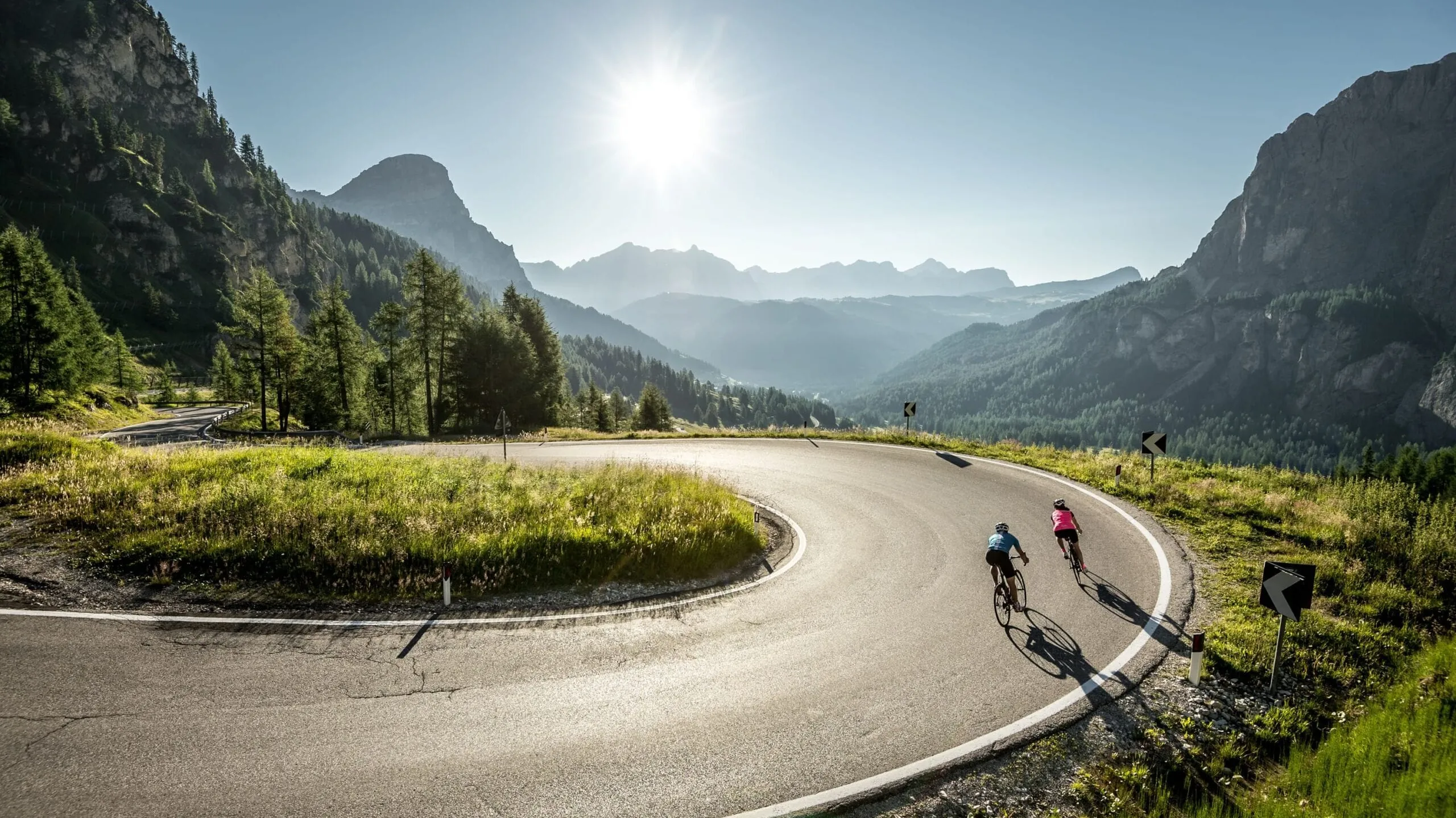 Curving Dolomites road with two cyclists riding uphill, sun over jagged peaks; pine trees on the left and grassy verges along the bend.