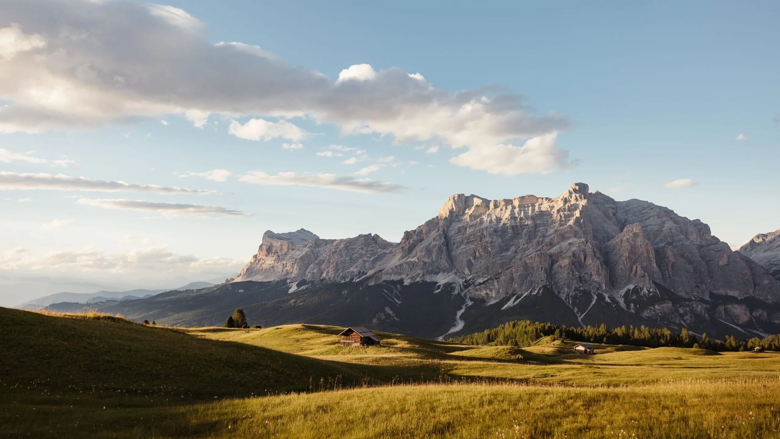 Sunlit alpine meadow in Alta Badia with a small wooden hut, rolling grassy hills, and towering Dolomites under a blue sky with scattered clouds.