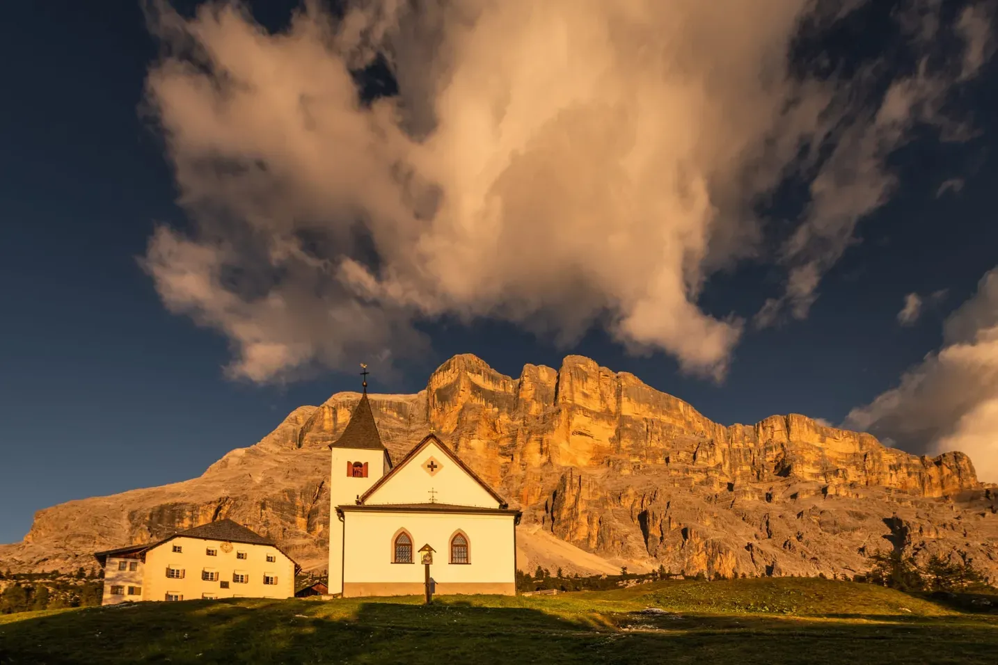 White church with a steeple on a grassy hill, Dolomites glowing gold behind under a blue sky with clouds in Alta Badia, Dolomites.