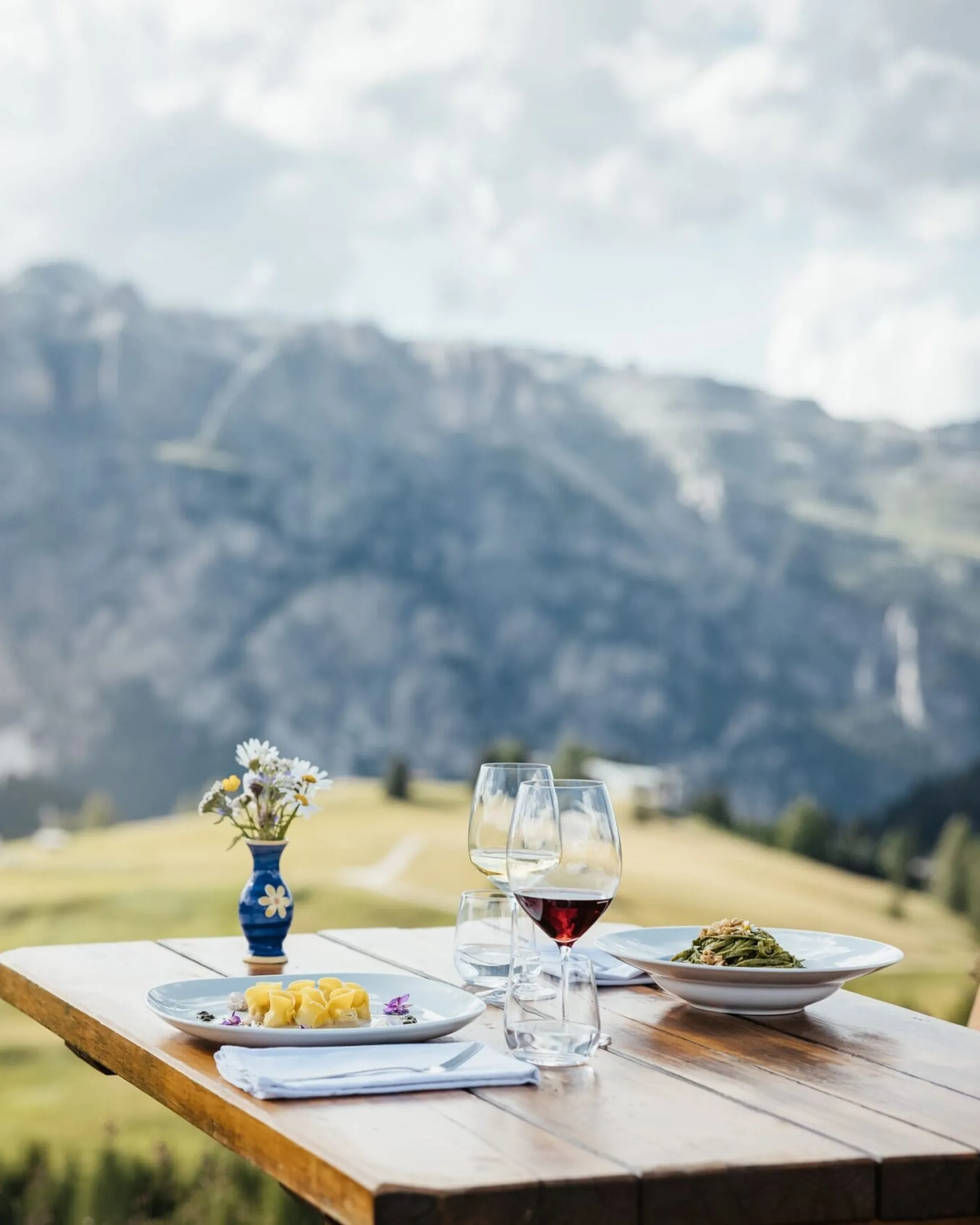 Tavolo in legno all'aperto con vista sulle Dolomiti: piatti, bicchieri di vino rosso e un bouquet di margherite in un vaso blu.
