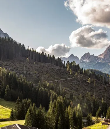 Paesaggio alpino in Alta Badia, Dolomiti: foreste di conifere, prati verdi e una strada serpeggiante che sale verso imponenti vette rocciose e nuvole.