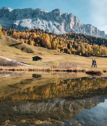 Autumnal Alta Badia landscape: golden trees, rugged Dolomites, a small cabin, and two people walking beside a calm lake that reflects the scene.