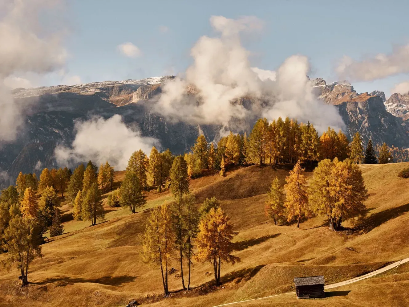Paesaggio autunnale in Alta Badia: pendii dorati da larici, una piccola baita di legno a destra, una strada e le Dolomiti avvolte tra nuvole sullo sfondo.
