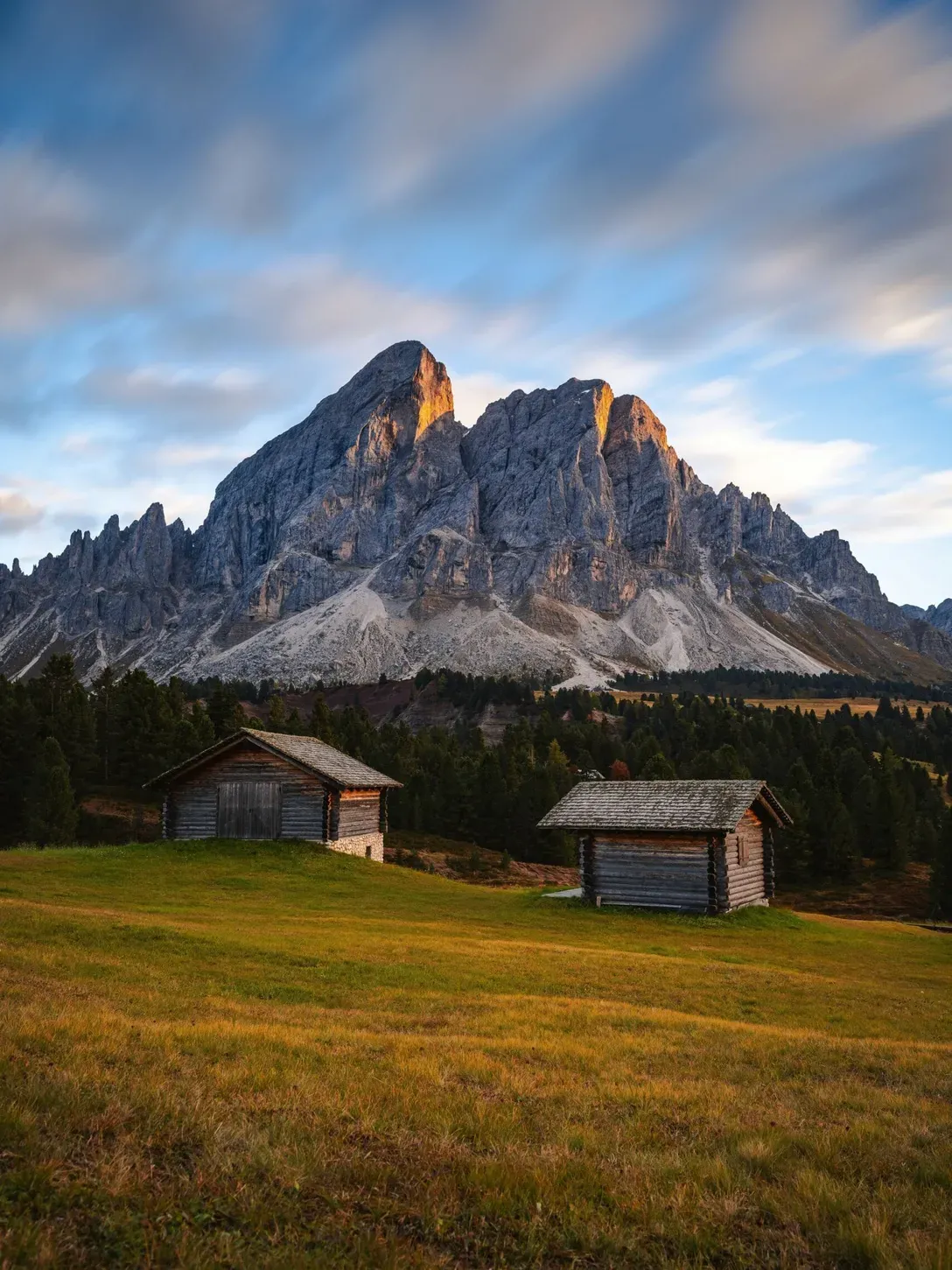 Due capanne di legno in un prato verde, con Dolomiti di Alta Badia sullo sfondo e cielo azzurro con nuvole, illuminato dal sole.