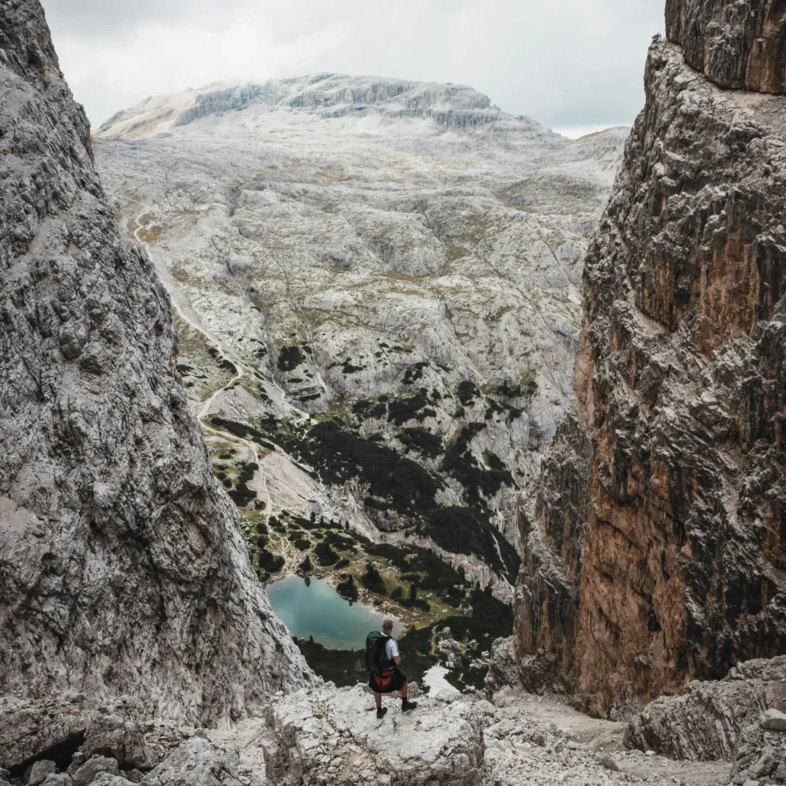 Una persona con zaino scende tra pareti rocciose maestose verso un lago turchese tra gole dolomitiche, in Alta Badia, Dolomiti.