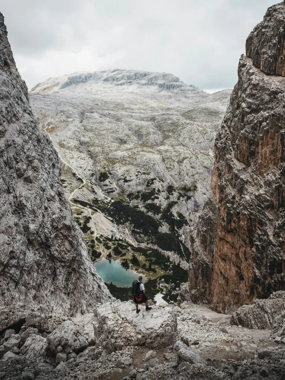 Hiker on a rocky Dolomite trail between tall cliffs, overlooking a turquoise alpine lake with rugged peaks in the distance.