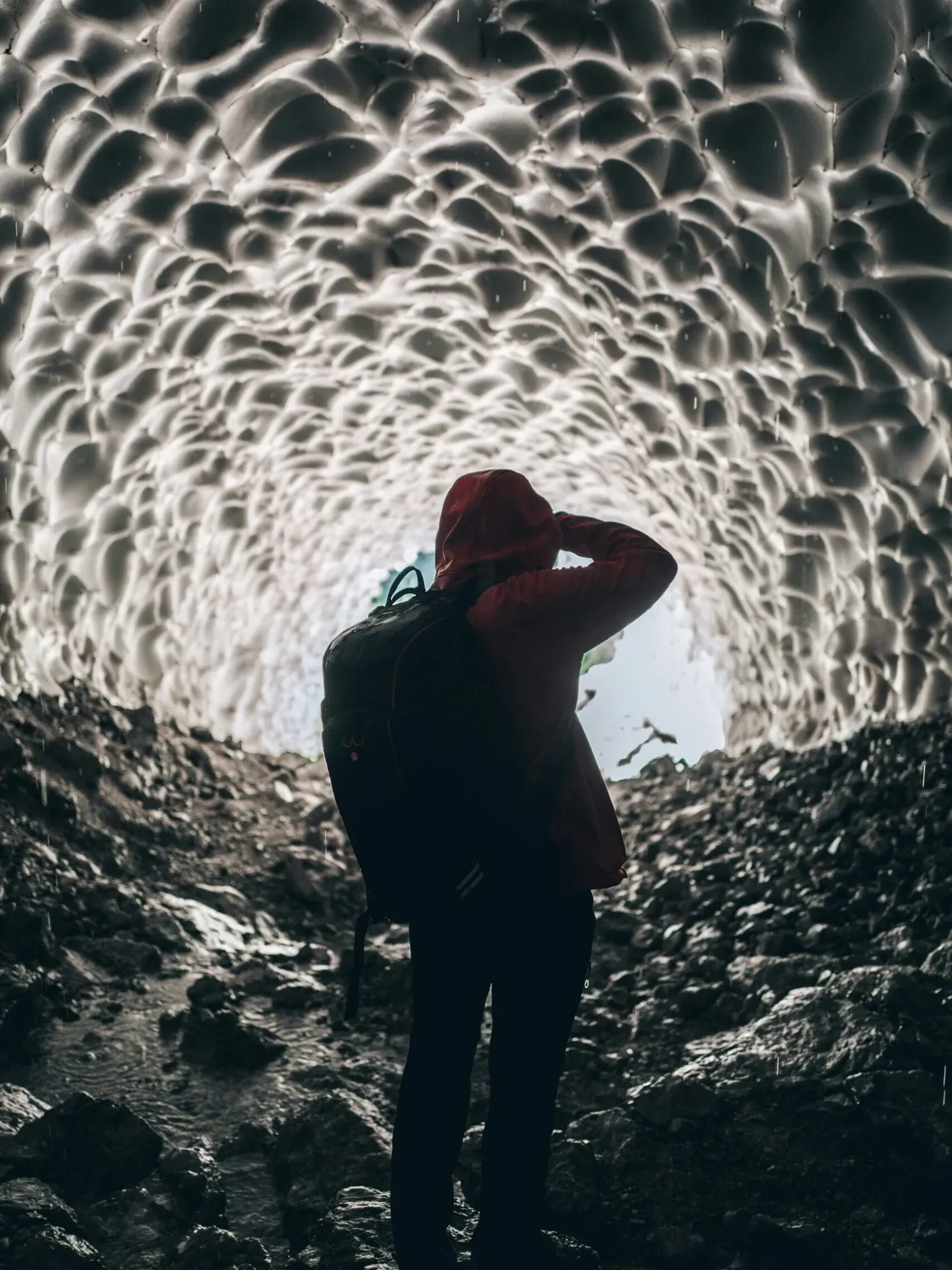 A hiker in a red jacket and backpack stands in a rocky ice tunnel with a honeycomb ceiling, looking toward the bright exit in the Dolomites.