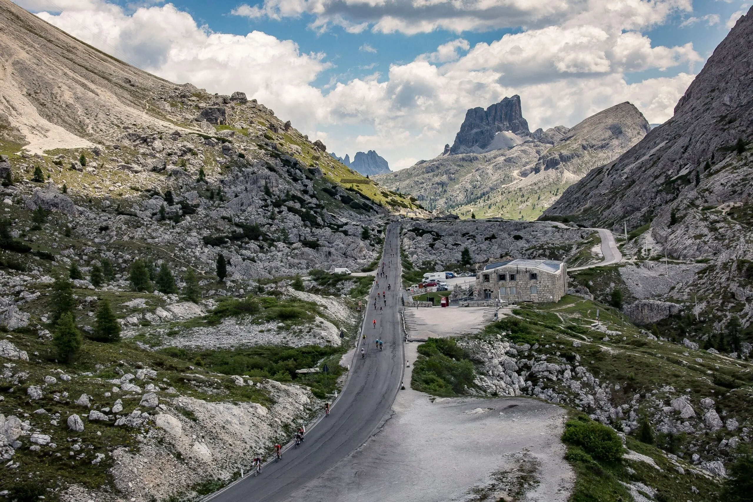 Winding mountain road through a rocky Dolomites valley with cyclists; a stone building sits beside the road as jagged peaks rise in the distance under a blue sky.