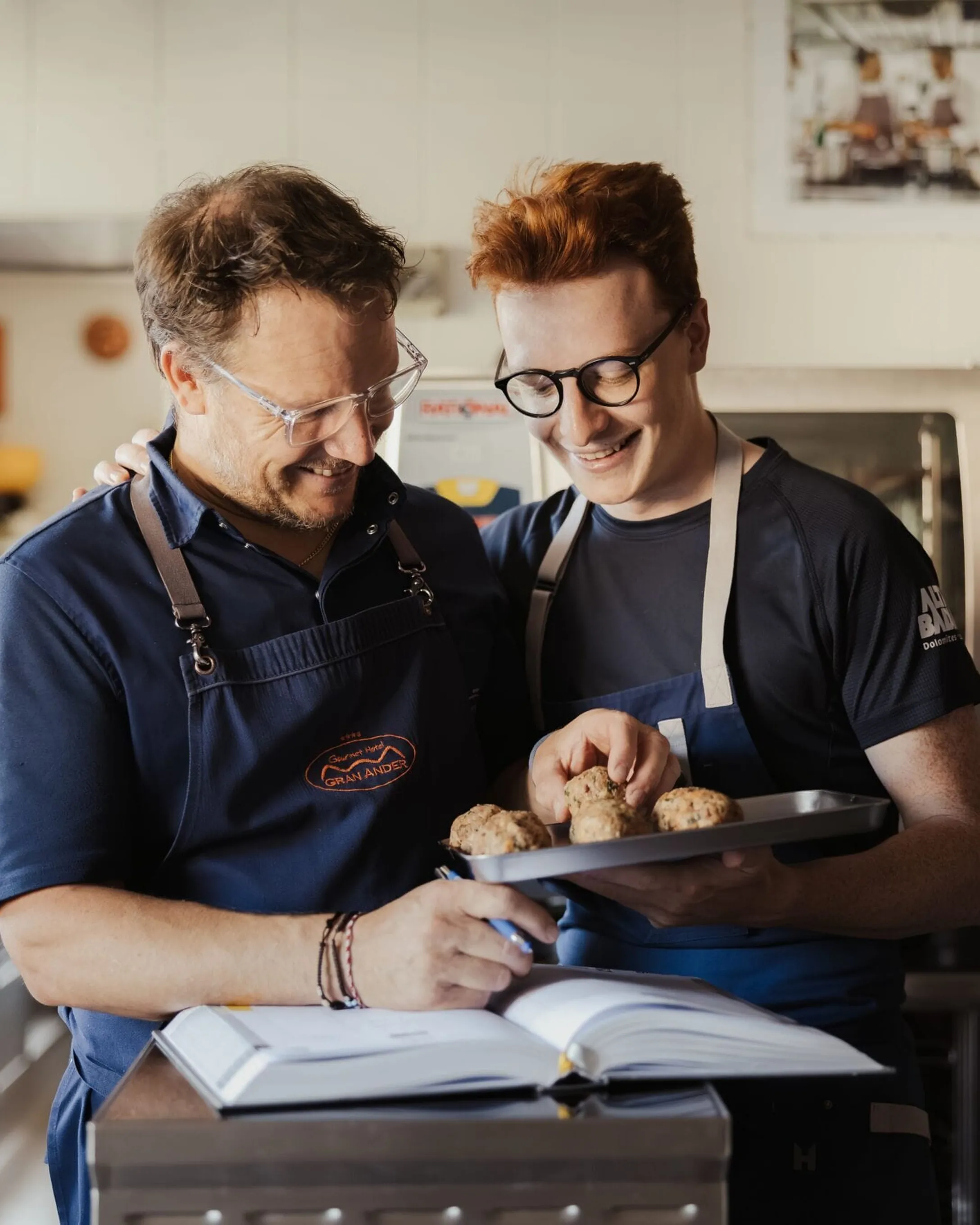 Maximilian e Andrea in cucina indossano grembiuli blu, sorridono mentre guardano un vassoio di dolcetti rotondi; sul bancone è aperto un libro di cucina.