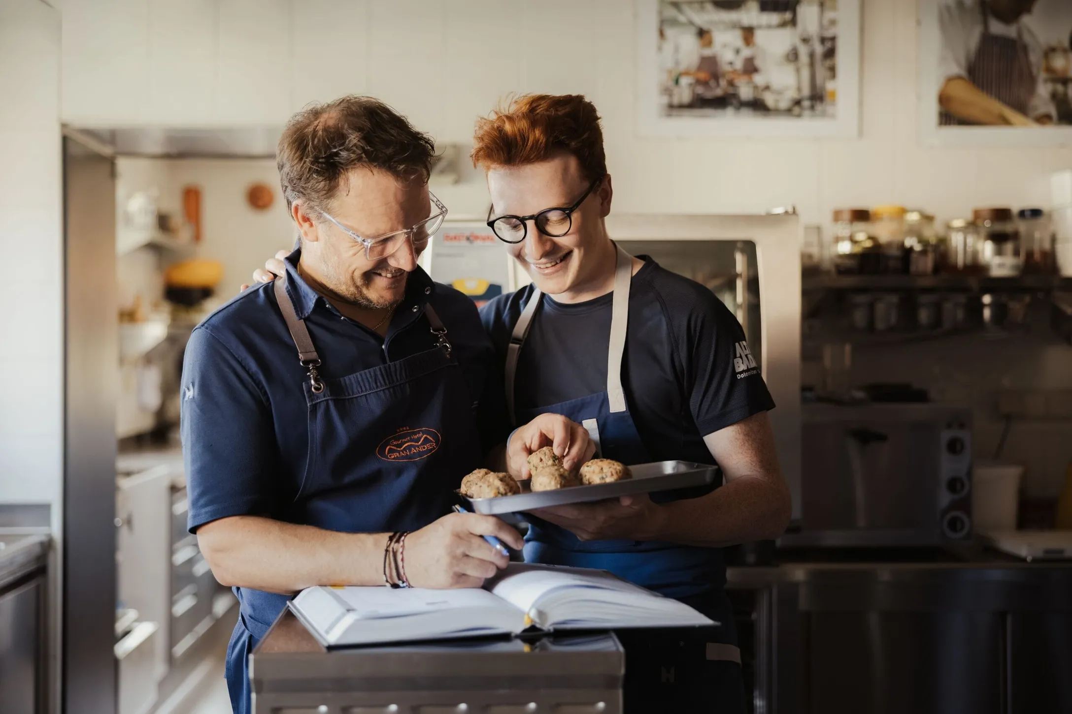 Maximilian and Andrea in the Hotel Gran Ander kitchen wearing blue aprons smile over muffins on a tray while referencing an open cookbook.