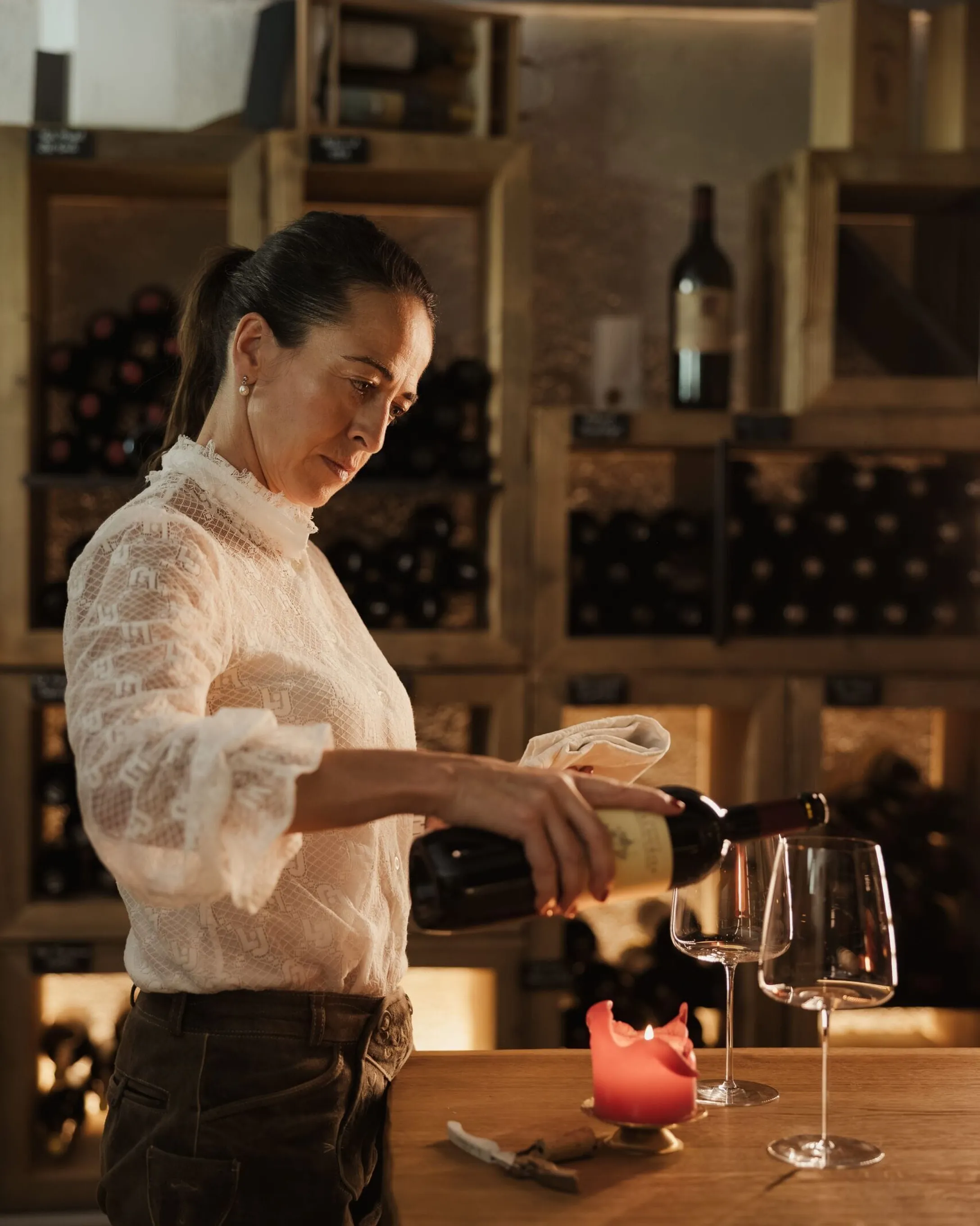 Evelin, wearing a white lace blouse, pours wine into a glass on the wooden counter; in the background, shelves full of bottles and a lit candle.