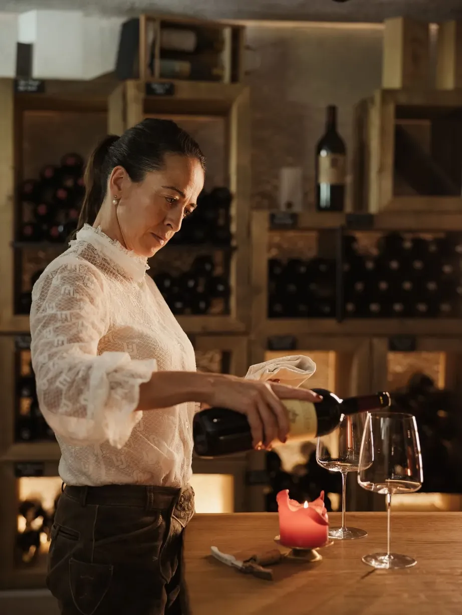 Evelin, wearing a white lace blouse, pours wine into a glass on the wooden counter; in the background, shelves full of bottles and a lit candle.