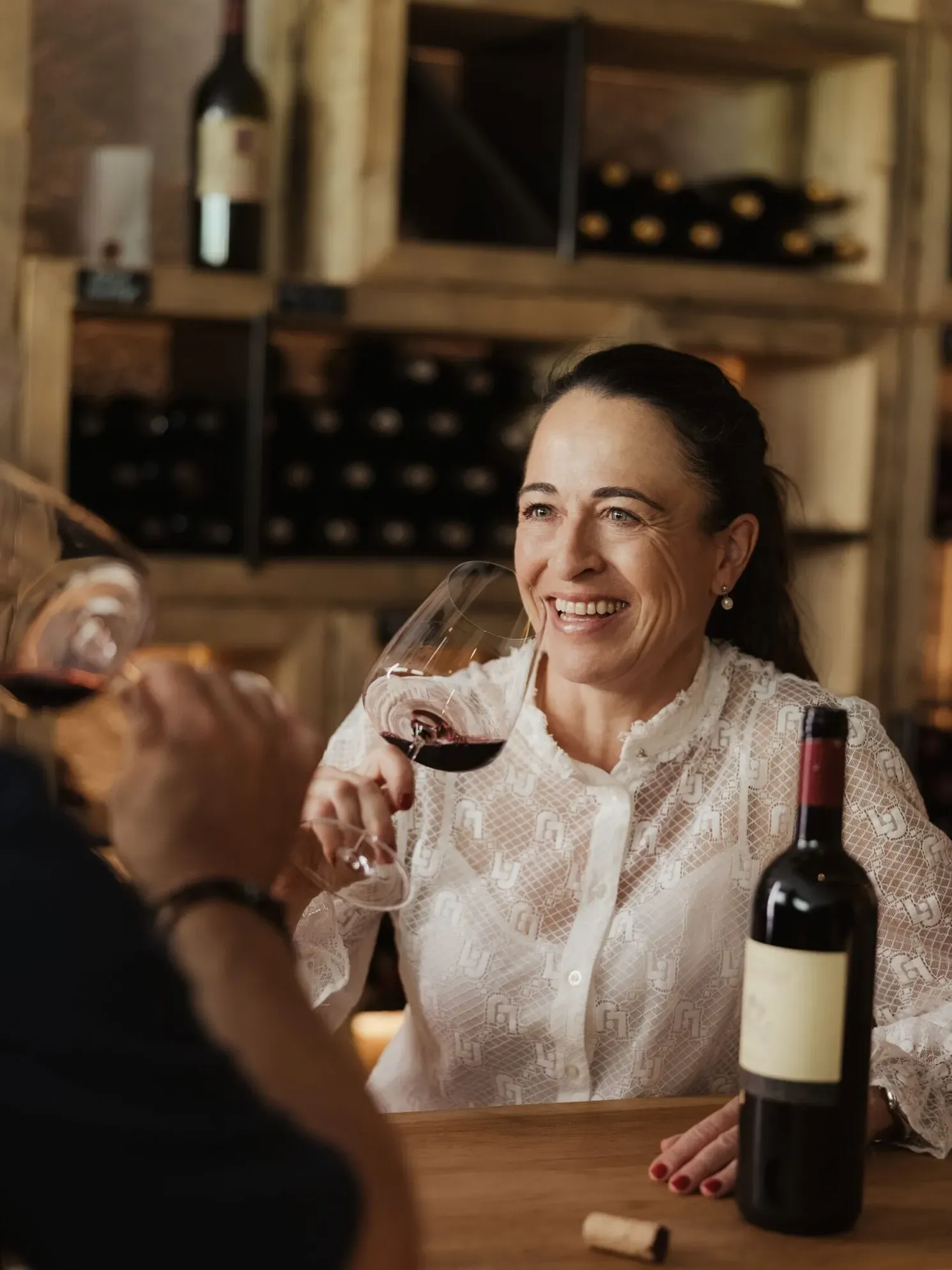 Evelin sits at the bar toasting with red wine; a bottle and cork are on the table, with shelves full of bottles in the background.