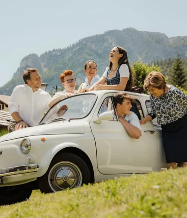 Famiglia Irsara su una Fiat 500 bianca d'epoca, felici sul prato a Alta Badia, Dolomiti, con le montagne sullo sfondo.