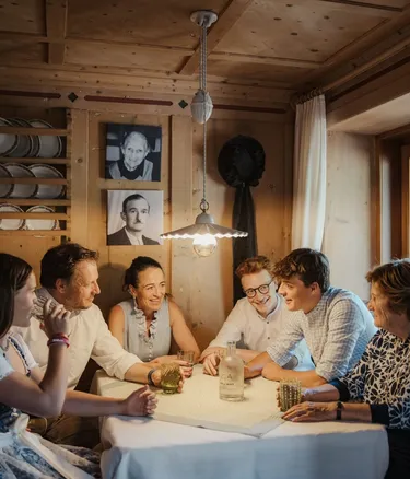 La famiglia Irsara attorno a un tavolo in una stanza rustica di legno, nell'Hotel Gran Ander, Dolomiti, illuminata da una lampada; bottiglia al centro e piatti appesi sul muro.