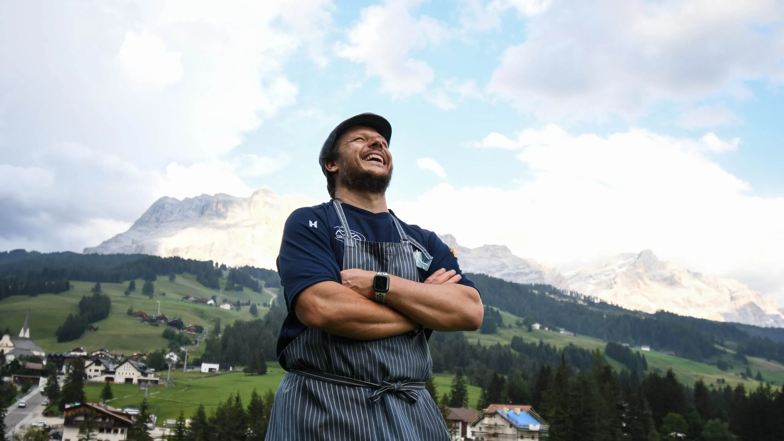 Andrea in grembiule a righe e cappellino sorride con le braccia incrociate, davanti alle Dolomiti e ai villaggi di Alta Badia.