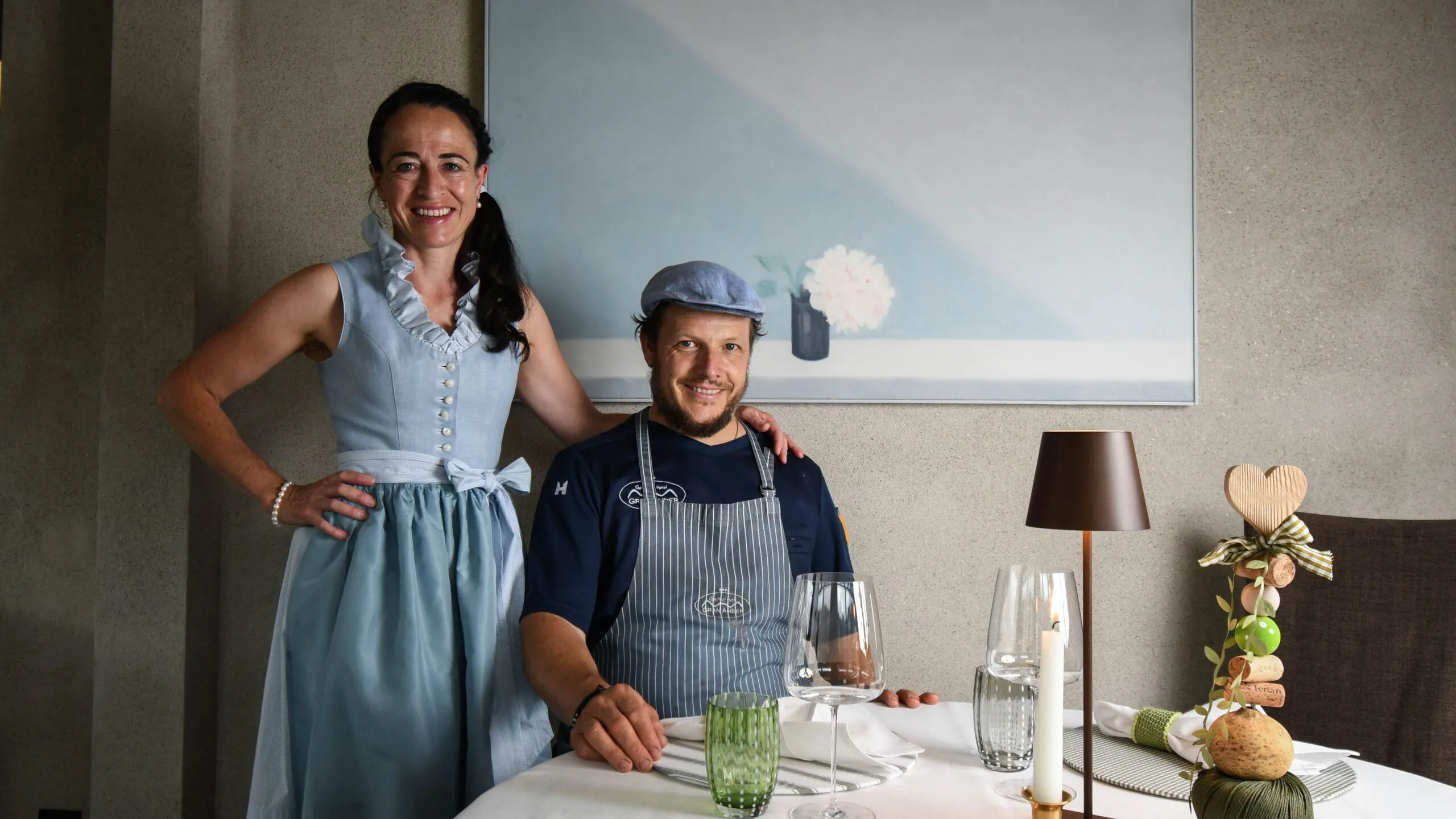 Evelin e Andrea sorridono a un tavolo nell'Hotel Gran Ander, Badia: una in vestito azzurro, l'altra in grembiule e berretto, con bicchieri e candela.