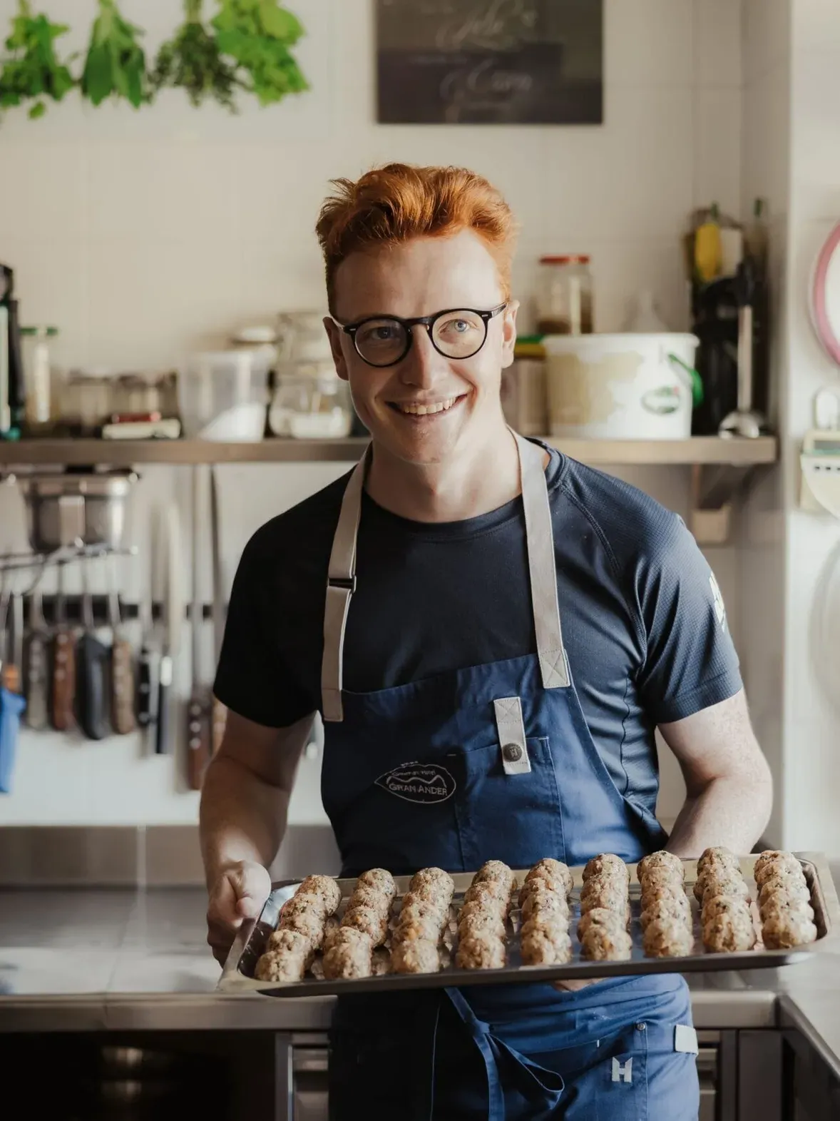 Maximilian wearing a blue apron holds a tray of baked cookies in a busy kitchen.