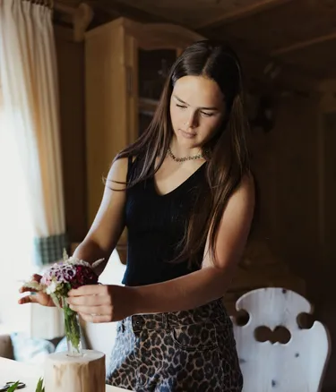 Lena con capelli lunghi scuri, top nero e gonna leopardata, sistema un bouquet in un vaso su un tavolo di legno.