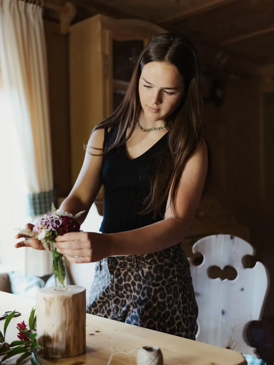 Lena, with long dark hair, a black top and a leopard-print skirt, arranges a bouquet in a vase on a wooden table.