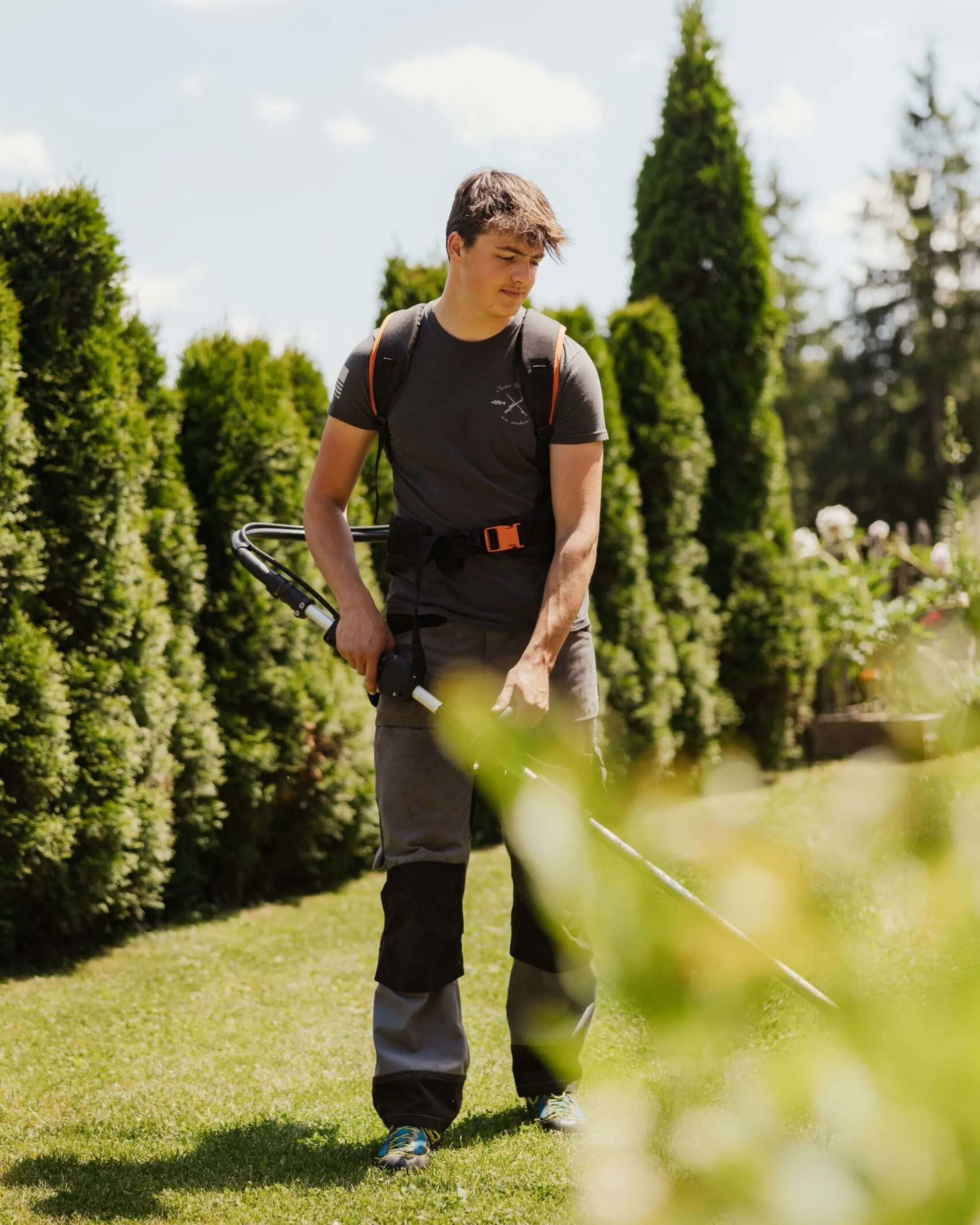 Tommy con zaino e abbigliamento da esterno usa un decespugliatore in un giardino soleggiato tra conifere in Alta Badia, Dolomiti.