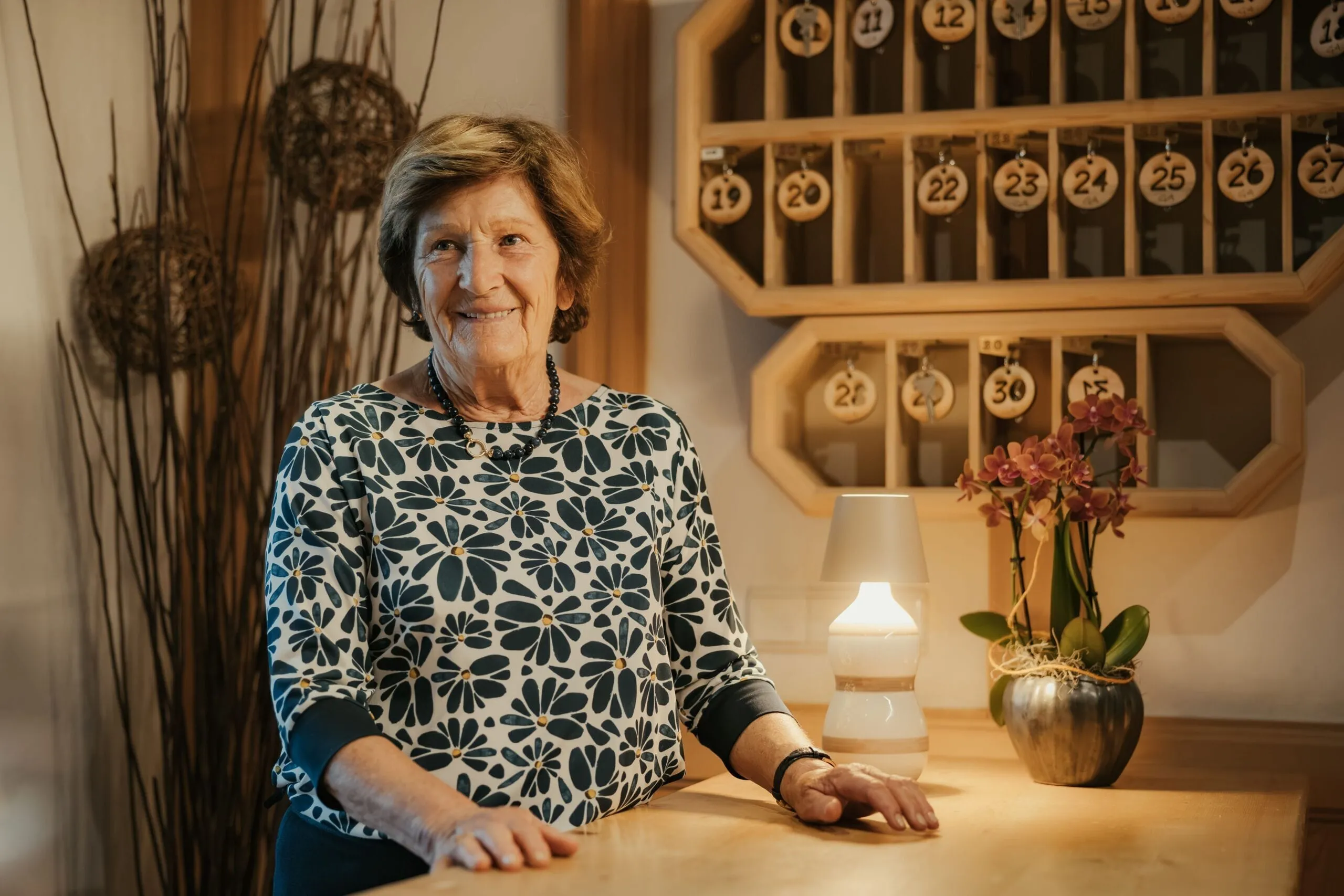 Rita smiling at the reception desk of the Hotel Gran Ander, Badia: wearing a blue and white floral shirt; against a background of a lamp and an orchid.