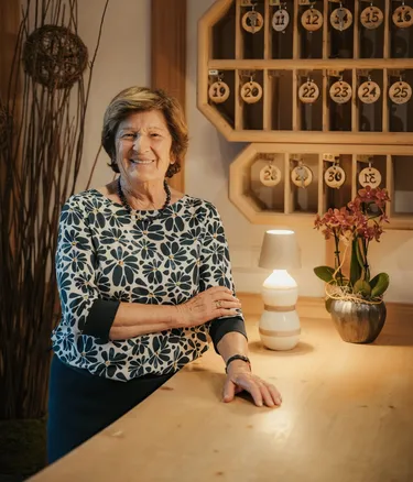 Rita sorridente dietro un bancone in legno nella hall dell'Hotel Gran Ander in Badia; sullo sfondo porta-chiavi in legno numerato, lampada accesa e vaso di orchidee.