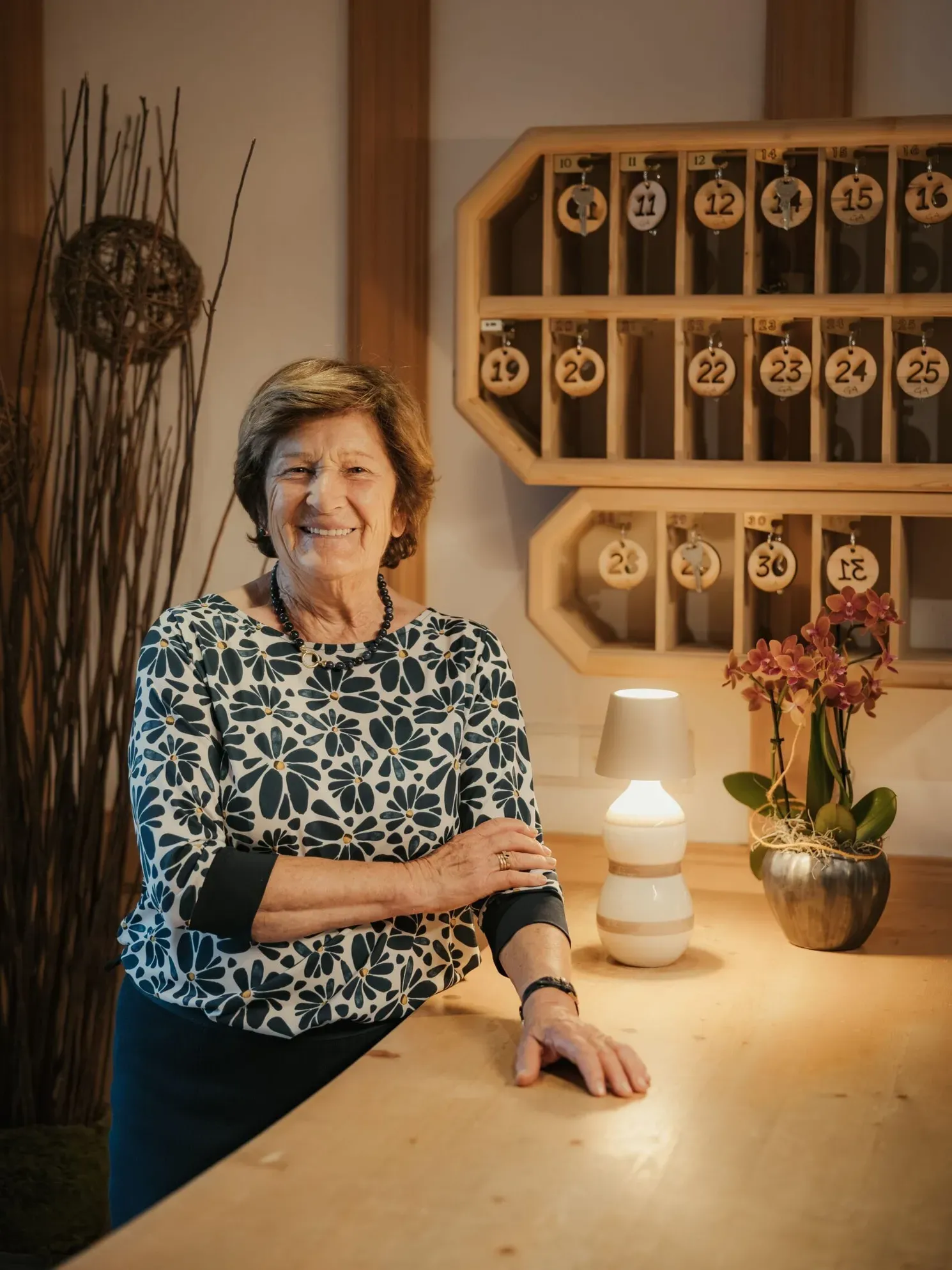 Inside Hotel Gran Ander, Rita with short hair in a blue floral blouse stands at a wooden reception counter, smiling beside a lamp and orchid.