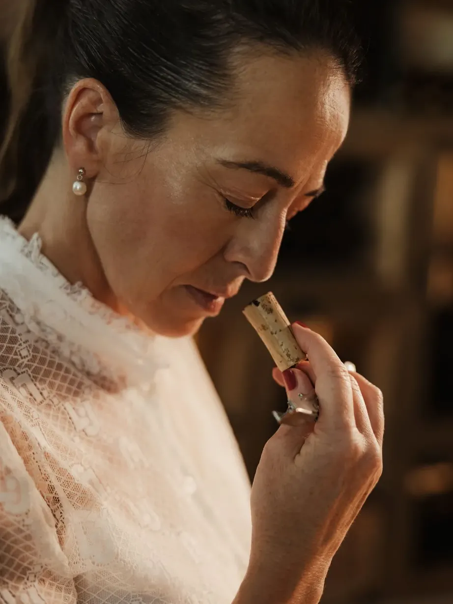 Evelin in foreground, left profile, hair tied back, pearl earrings, white lace top, holds a small cosmetic tube to her lips.