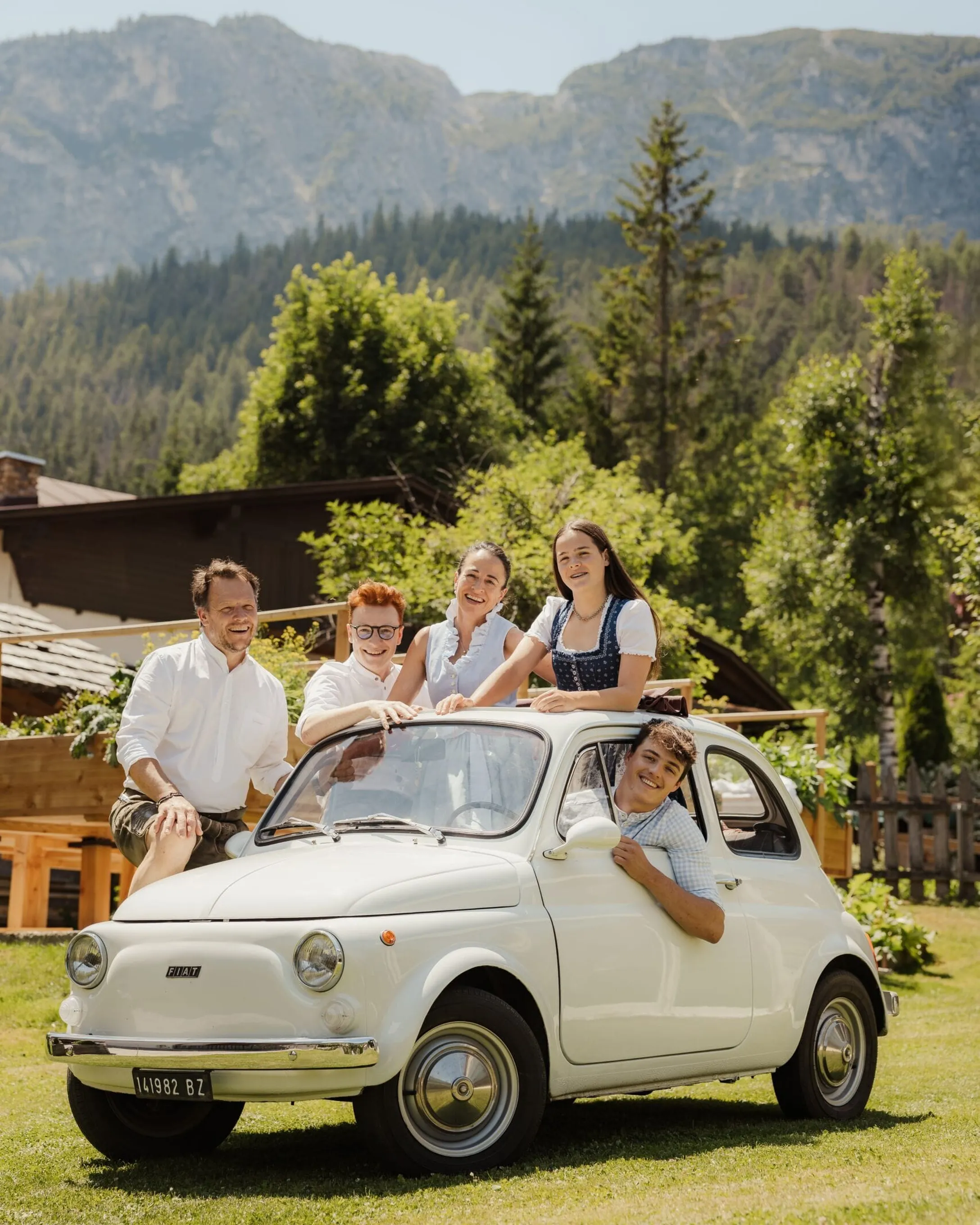 I componenti della famiglia Irsara sorridenti si appoggiano a una Fiat 500 bianca d'epoca in un prato verde, con case e il paesaggio delle Dolomiti sullo sfondo.