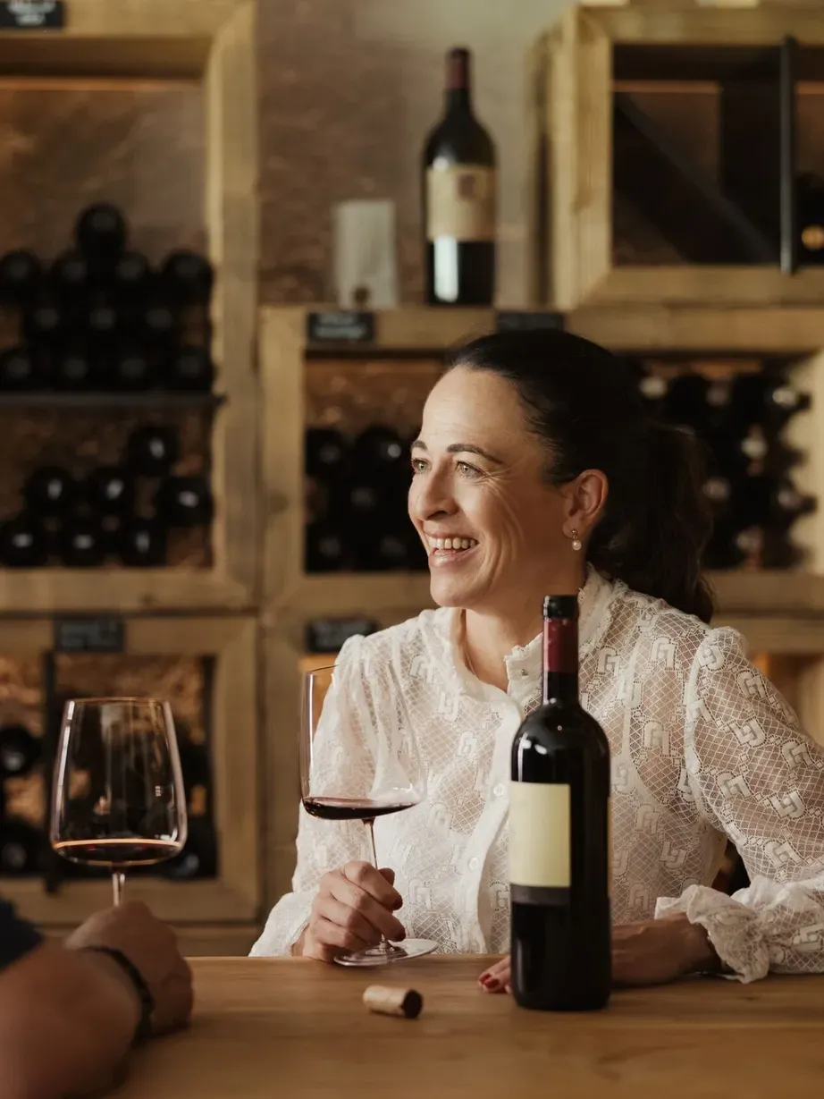 Evelin and Andrea at the wine cellar counter: one person smiles with a glass of red wine; on the table, a bottle and a cork, against a backdrop of shelves full of bottles.